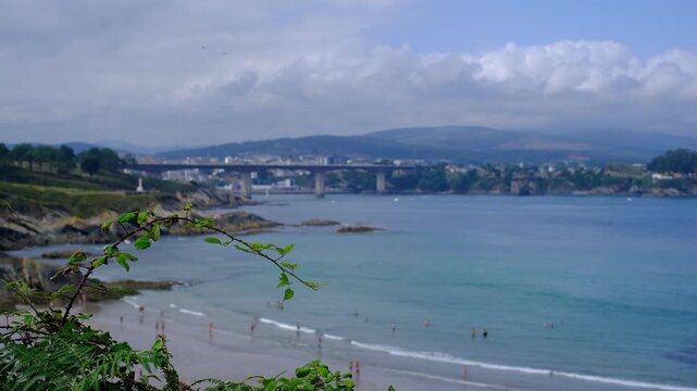 Scenic view of Playa de Arnao in Castropol, Asturias, featuring a picturesque sandy beach along the calm blue estuary waters, framed by lush coastal greenery and a distant bridge under a cloudy sky
