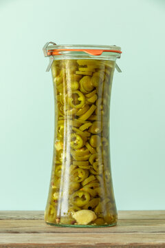 Asian style pickled jalape&ntilde;o slices in a glass jar on a rustic wooden table. Spicy pepper rings in homemade sweet-and-sour brine, close-up on a pale green pastel background with natural side light.