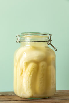 Lacto-fermented White Wonder cucumbers: sour pickles in a glass jar. Organic homemade heritage-variety gherkin pantry preserves on a rustic table with a pastel-green background. 
