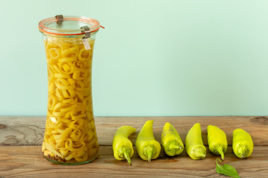 Hungarian Hot Wax pepper slices in sweet and sour brine jar. Asian pickled chili rings with six whole peppers row on rustic wood table close up on pale green pastel background with copy space.