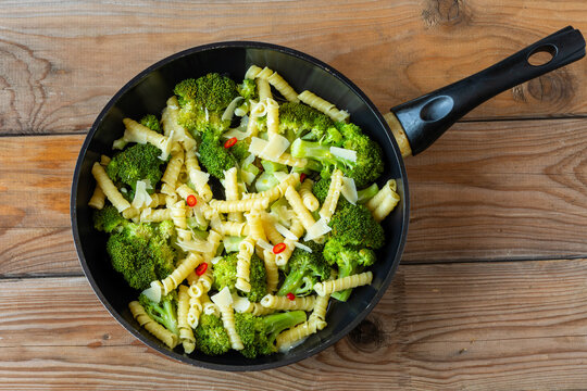 Steamed broccoli with riccioli pasta, garlic, and parmesan in a frying pan. Italian homemade savory noodle dish with cheese, top view on a rustic wooden background.