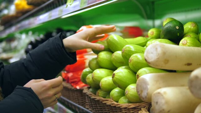 Female customer hands selecting fresh green zucchini from a basket in a supermarket produce section. Woman shopping for healthy organic vegetables for a balanced and nutritious diet