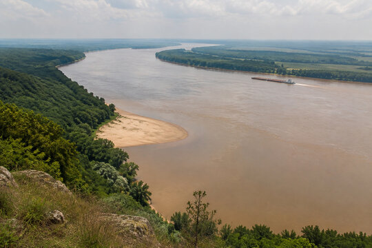 View from high bluff of wide brown river bending through low green forest, sandbar, tugboat and barge downstream, Mississippi River landscape