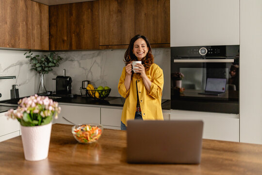 Young woman enjoying coffee in modern kitchen while working on laptop and smiling