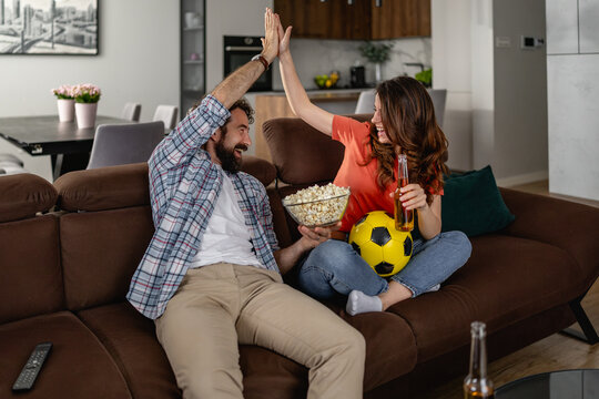 Couple on sofa high-fiving while watching soccer with popcorn, beer and a soccer ball