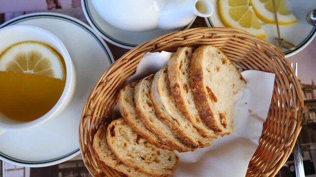 Top view of a female hand taking a freshly sliced piece of rustic sourdough bread from a wicker basket, next to a cup of lemon tea, creating a cozy and warm breakfast scene