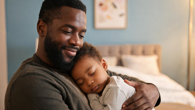 A caring father holds his sleeping young child closely in a cozy bedroom, showing tender affection.