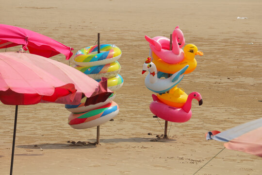 Colorful inflatable swim rings in animal shapes like flamingo and duck on a sandy beach with pink umbrellas.