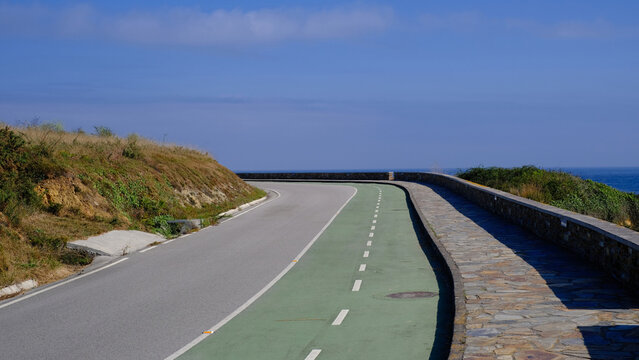 Scenic view of a coastal road and dedicated green bike lane curving along a stone wall overlooking the deep blue sea under a clear sky, capturing a peaceful and picturesque cycling route for travel an