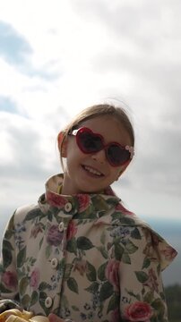 Child portrait happy girl in sunglasses smiling against a cloudy sky on a mountaintop.