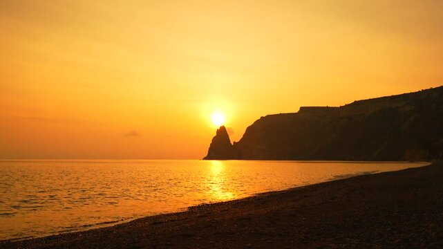 Sunset, sea stack, beach. Incredible golden sun sets dramatically behind a rugged rocky cliff overlooking the ocean waves.