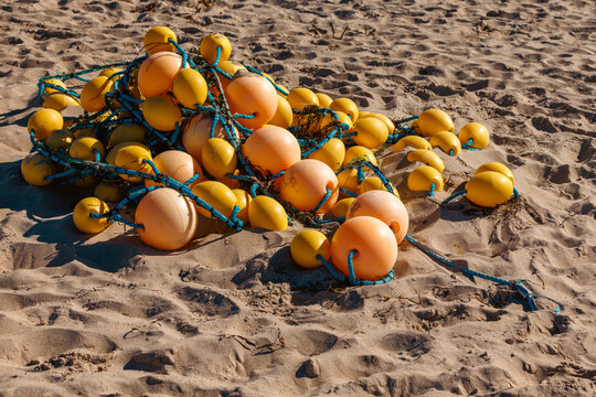 Large cluster of orange and yellow fishing buoys with blue ropes resting on a textured sandy beach in sunlight.