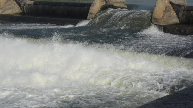 Wide slow motion view of strong river water discharging over an inflatable rubber dam structure with small birds flying across the foaming white current.
