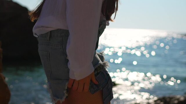 Woman beach fashion wearing denim shorts over a plaid kilt by the shimmering blue sea during a bright sunny summer day