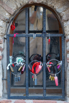 A close-up view of an arched window with a black iron security grate decorated with colorful love padlocks attached to its rings, set within an ancient stone structure