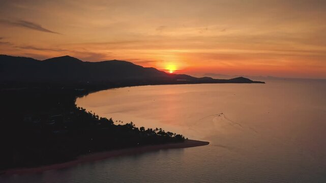 Warm golden sunset over calm ocean, reflecting on water, mountains and tropical beach coastline in background, boats leave trails on the surface. Travel Samui tropical island. Aerial view drone flight