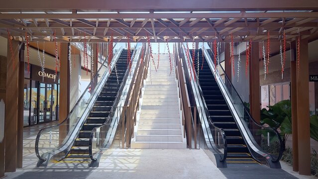 Symmetrical architectural view of modern outdoor escalators and a central staircase inside a tropical shopping mall, featuring a wooden canopy, glass railings, and hanging festive decorations