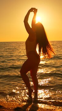 Ocean sunset silhouette of a woman forming a heart shape with hands while standing in shallow water. Static wide shot.