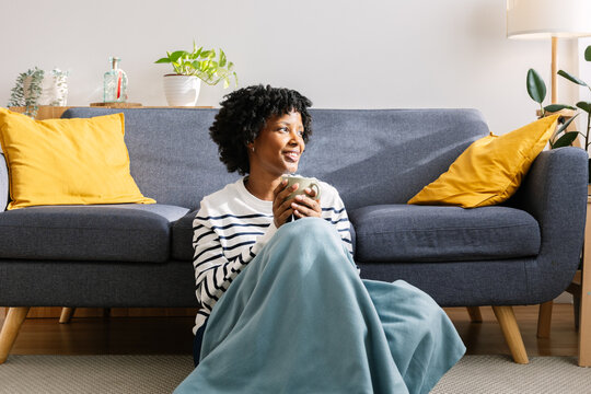 girl sitting in living room with cup of hot coffee enjoying under blanket. Joyful black girl sitting on floor by the sofa wrapped in blanket in living room with cup of hot coffee