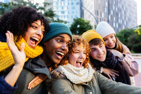 Five cheerful multi-ethnic young students are taking a selfie in the city during winter, wearing wool hats and scarves