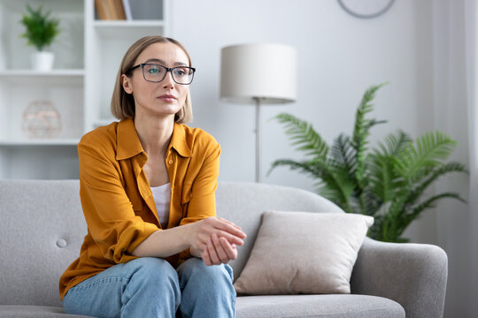 Thoughtful woman feeling sad and worried, sitting alone on a sofa in a modern living room, contemplating problems and experiencing anxiety, loneliness, and depression