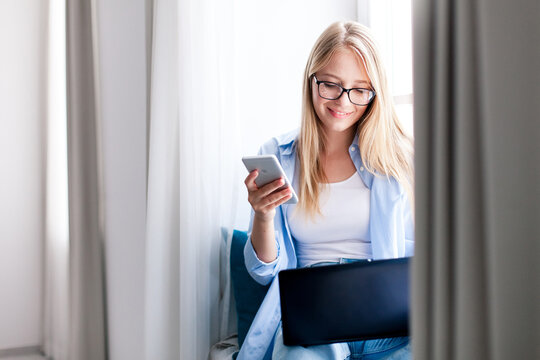 Young woman working from home office. Happy freelancer using laptop and mobile phone. Workplace in living room on windowsill. Concept of female business, teleworking, wellbeing