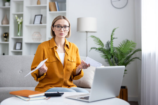 Woman on sofa reviewing bills and paperwork with laptop and calculator on table, looking worried and overwhelmed while managing household finances and budgeting at home