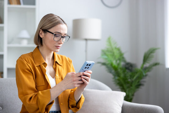 Woman in eyeglasses sitting on a sofa, concentrating on her mobile phone, reading a message with a focused and serious expression, experiencing online communication