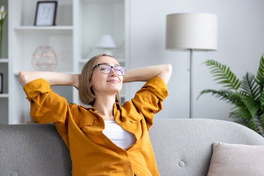 Woman in eyeglasses sitting on a sofa with hands behind her head, eyes closed, enjoying a moment of calm and mindfulness in her comfortable modern living room
