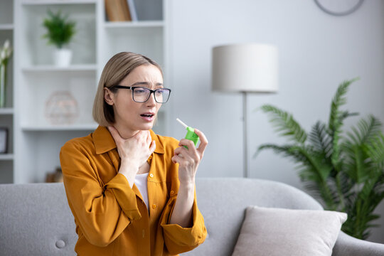 Young woman feeling discomfort from a sore throat and holding a medicated throat spray while sitting on a sofa in a bright home living room, seeking immediate relief from the illness