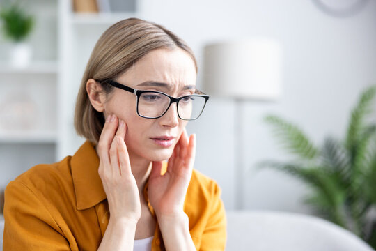Mature woman in glasses touching her cheek and wincing from severe tooth or jaw pain, distressed and uncomfortable indoors at a dental clinic or medical appointment, close-up
