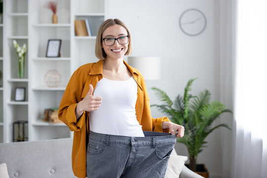 Young woman smiling, holding her oversized old jeans, and giving a thumbs up, representing her successful weight loss journey and body transformation at home