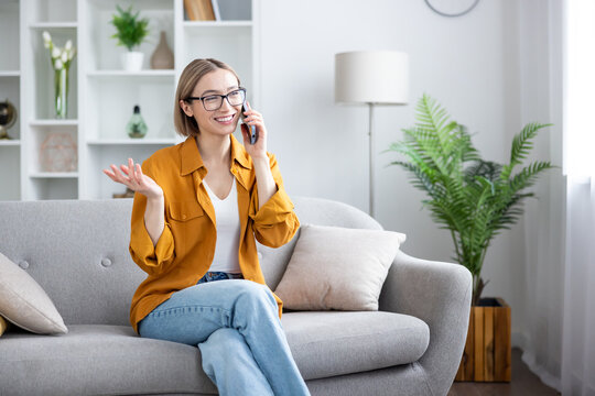 Young woman wearing glasses and a yellow shirt, sitting comfortably on a gray sofa in a modern living room, smiling and talking on her smartphone while gesturing with her hand