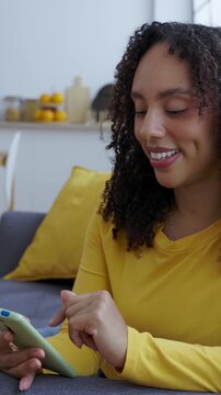 Vertical HD video of young African American woman scrolling on smartphone while relaxing at home. Smiling female enjoying social media content on the sofa in a modern apartment