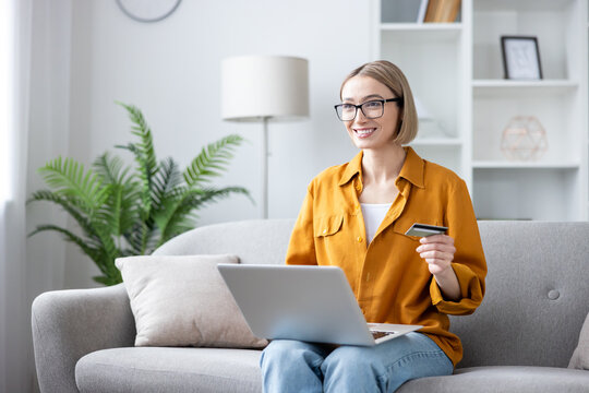 Young woman wearing glasses and a yellow shirt sitting comfortably on a gray sofa, smiling happily while using her laptop and holding a credit card for convenient online shopping at home