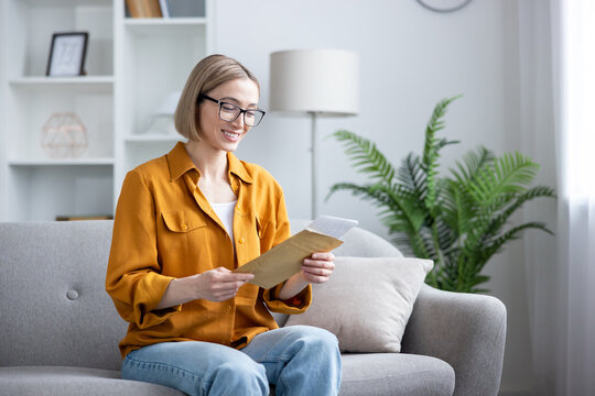 Young woman on a cozy modern sofa, smiling as she opens a brown envelope and reads an important letter, receiving positive news and feeling surprised and relieved