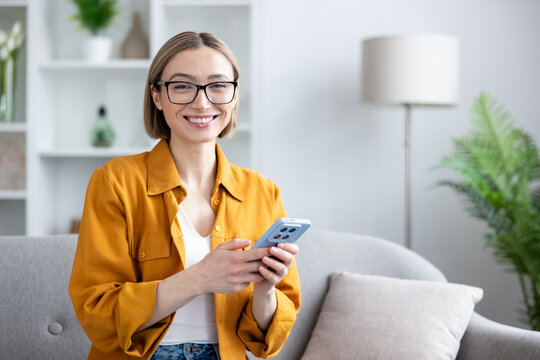 Young woman wearing eyeglasses sitting on a sofa at home, smiling at the camera and holding a mobile phone, representing modern communication, technology, and a relaxed lifestyle