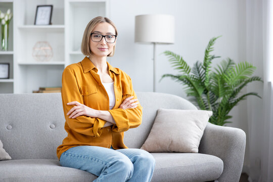Young woman wearing glasses and a yellow shirt, sitting confidently on a gray sofa at home, crossing her arms while looking at the camera, conveying professionalism and relaxation