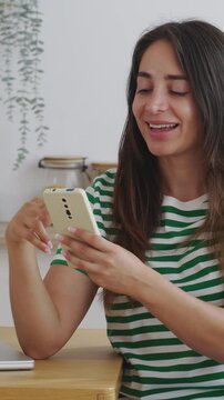 Vertical HD video of beautiful young Latin American woman with long hair sitting at her kitchen table, smiling and laughing while scrolling through her social media feed on a modern smartphone
