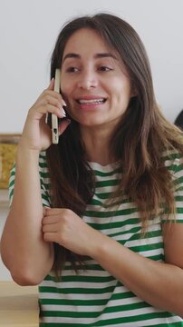 Vertical HD video of young adult happy Latin American woman talking on phone call with friend standing in the kitchen at home. Communication and technology lifestyle concept