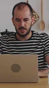 Vertical HD video of focused mid adult man sitting at his kitchen table and using a laptop for remote work, looking at the screen with concentration while thinking about a project. Business concept
