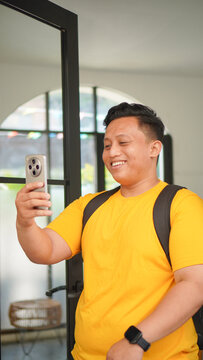 Young Asian man in a yellow shirt making a video call on his smartphone, smiling and waving his hand near a glass door indoors. Casual lifestyle and online communication concept, vertical format.