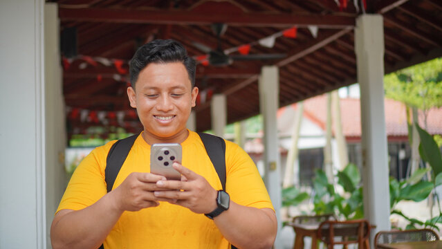 Happy young Asian man in a yellow shirt smiling while looking at his smartphone, wearing a backpack and smartwatch under a wooden pavilion outdoors. Casual lifestyle and mobile technology concept.