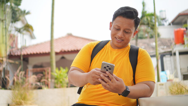 Young Asian man in a yellow shirt holding a smartphone with both hands, smiling and looking up thoughtfully outdoors. Casual lifestyle concept of a student or traveler using mobile technology.