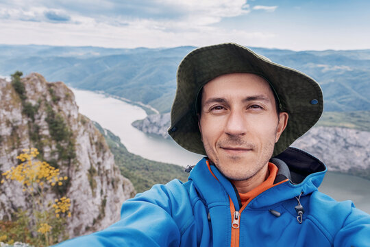 Close-up selfie of a smiling male hiker in a sun hat framing a dramatic Danube river gorge and mountain panorama behind him. Adventure travel concept.