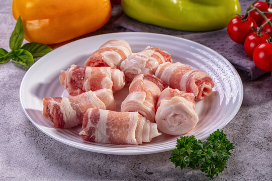 Raw bacon wrapped meat rolls on white plate with bell peppers and tomatoes