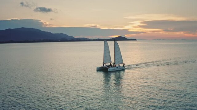 Samui, Thailand - 02.16.2022: Catamaran sailing on the calm ocean water, leaving a wake behind, with a beautiful sunset sky and distant islands, creating a serene and idyllic tropical scene. People