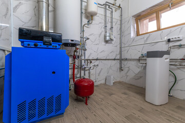 A blue boiler stands in a utility room with metal pipes and a red water tank. The walls have white tiles, and there are windows letting in natural light © Alhim