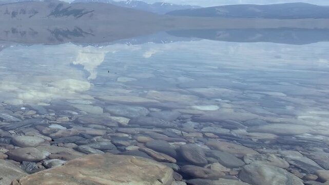 Discover the ethereal clarity of Lake Khuvsgul in Mongolia. Sunlight illuminates the rocky lakebed through the pristine water, mirroring majestic mountains and sky above. A tranquil natural wonder.