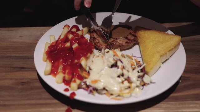 Person cutting grilled pork steak with knife and fork on white plate. Served with french fries with ketchup, fresh coleslaw, and toasted bread on wooden table. Delicious restaurant meal setup.